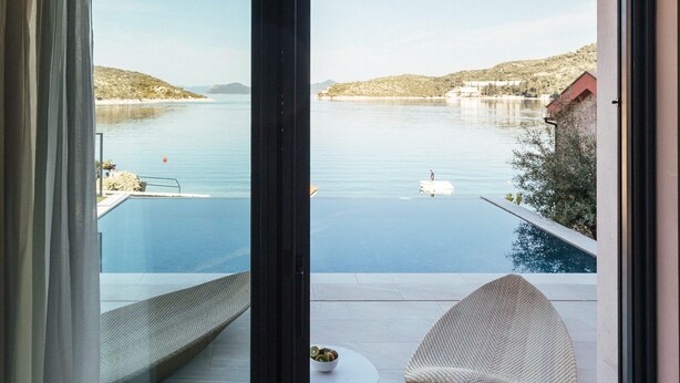 Habitación con un amplio ventanal corredero que da a una piscina con vistas a un lago y colinas. Hay una silla y una mesita en la habitación.