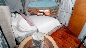 A cozy tented room with a bed, a table with a mug, and a view of a wooden deck and mountains.