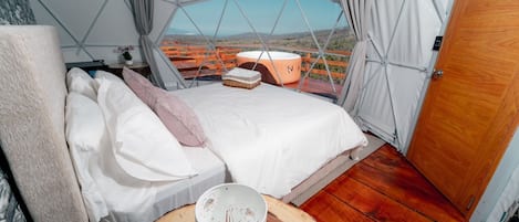 Une pièce accueillante aménagée sous une tente, avec un lit, une table et une tasse, et une vue sur une terrasse en bois et les montagnes.
