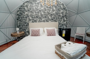 Una cama con una pared estampada en piedra, una mesita de noche con una lámpara, y una cesta de toallas.