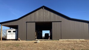 A large, dark metal barn with an open door, a white trailer parked outside, and a dog standing in front.