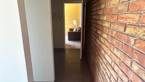 Ein Korridor mit einer Backsteinwand auf einer Seite und einer weißen Wand auf der anderen, der zu einem Raum mit einem Ventilator und einem Stuhl führt.