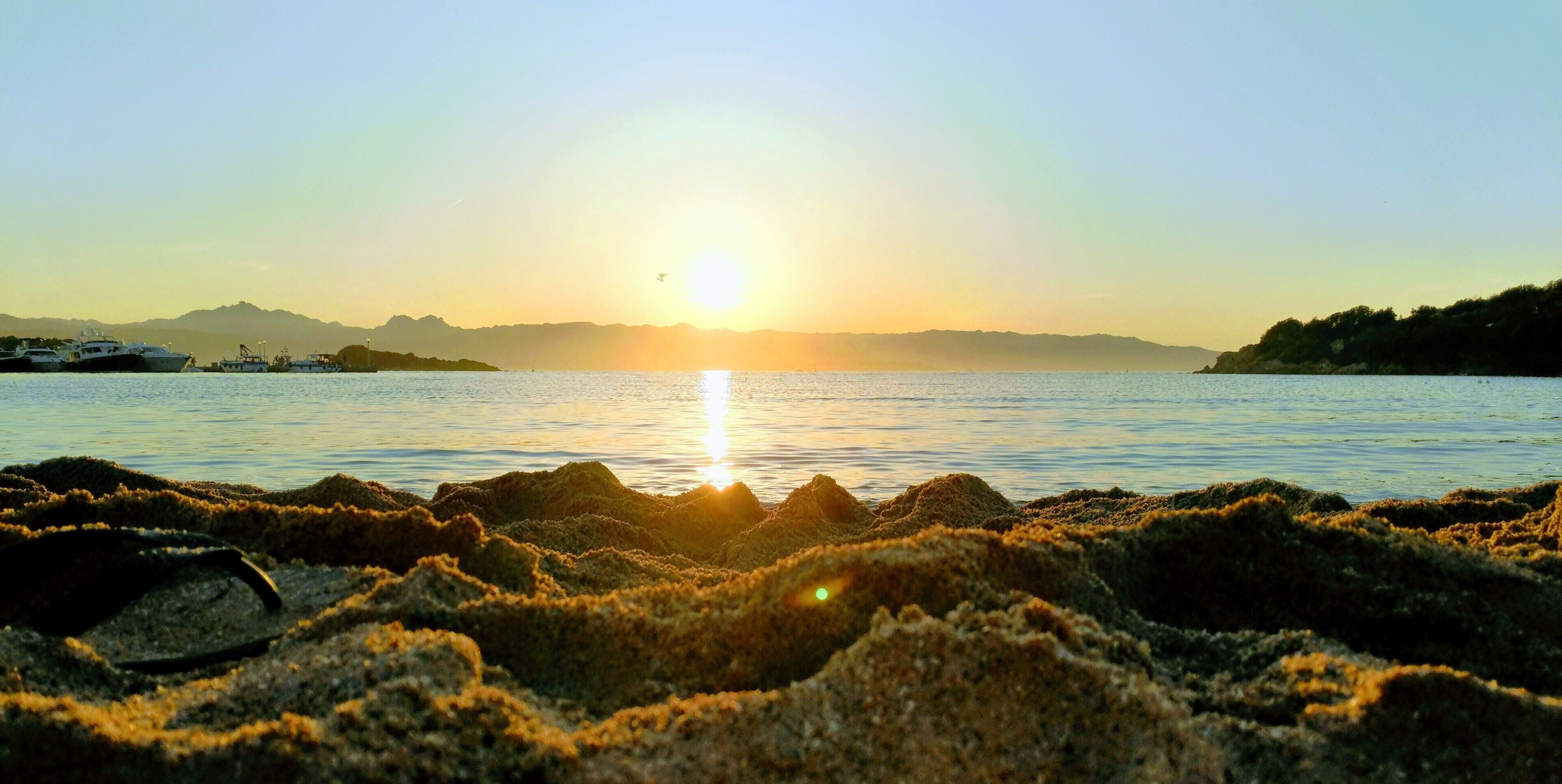 A sunset over a body of water with boats and rocky terrain in the foreground.