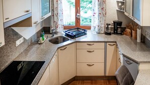 Eine kleine Küche mit Spüle, Herd und Schränken. Ein Fenster mit blumigen Vorhängen und eine Kaffeemaschine auf der Arbeitsplatte.