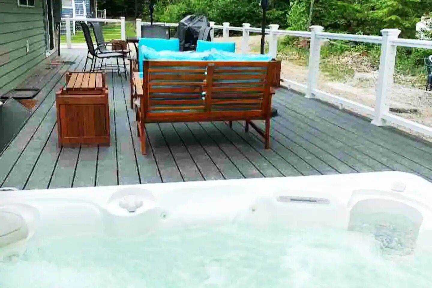 Una terraza de madera con jacuzzi, mobiliario exterior y una valla blanca.
