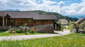 Un bâtiment traditionnel en bois, doté d’un mur en pierre et d’une clôture en bois, situé dans un paysage rural, avec un ciel dégagé et des montagnes en arrière-plan.