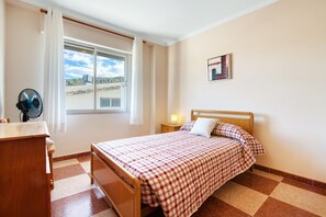 A bedroom with a checkered bedspread, a wooden bedside table, a nightstand with a lamp, a window with curtains, and a fan. - Apartment 'La Bodegueta' with Private Terrace, Wi-Fi and Air Conditioning (Orba)