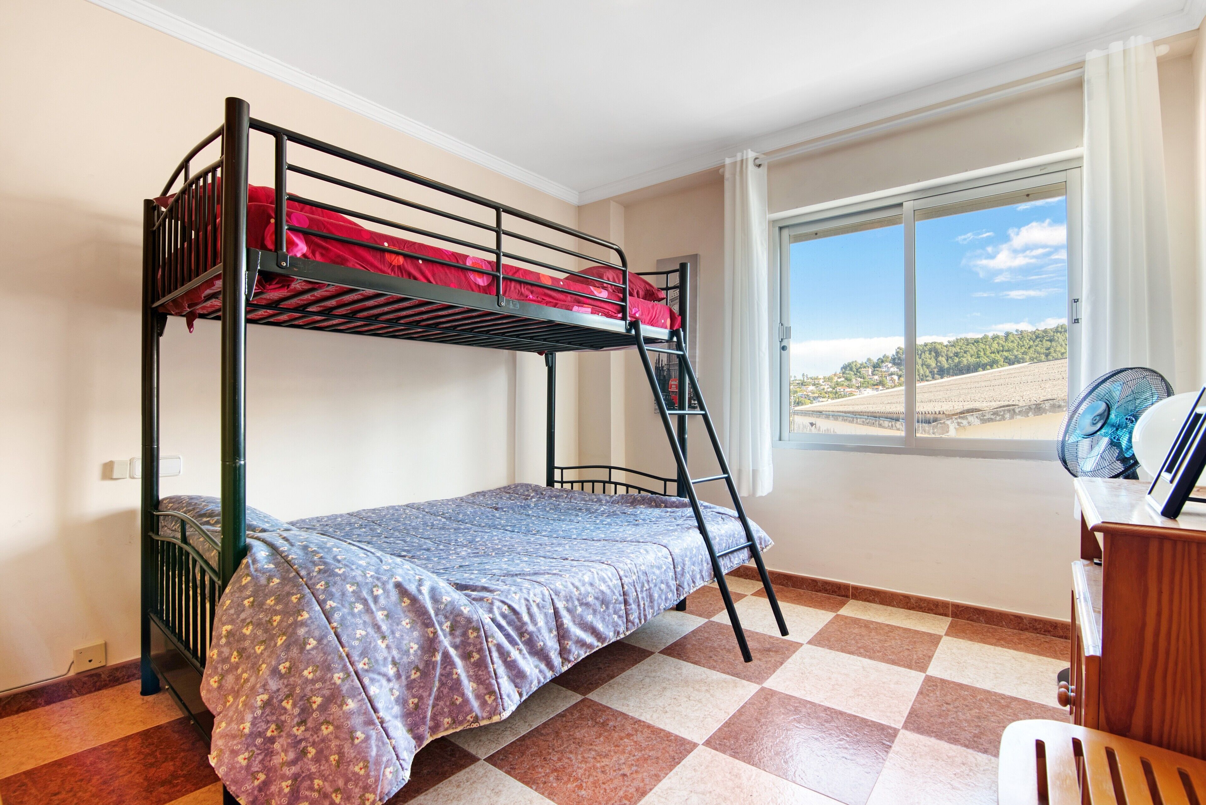 Une chambre avec un lit superposé, un bureau avec une chaise, une fenêtre donnant sur un paysage agréable et un ventilateur.