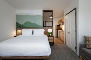 Habitación de hotel con una cama grande, mesitas de noche con lámparas, una kitchenette pequeña y un baño visible a través de una puerta.