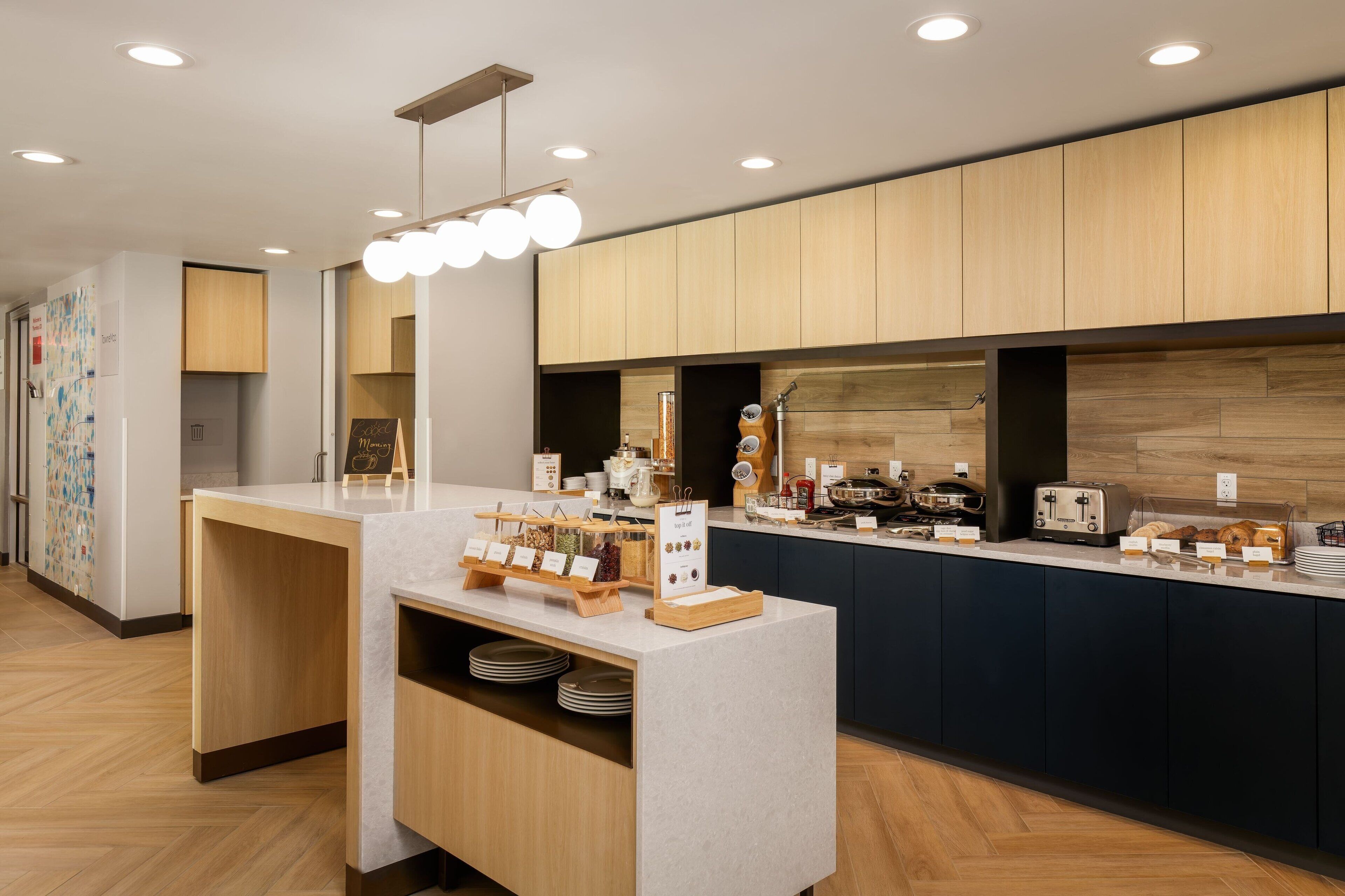 A modern hotel kitchen with wooden cabinets, a marble countertop, and various breakfast items.