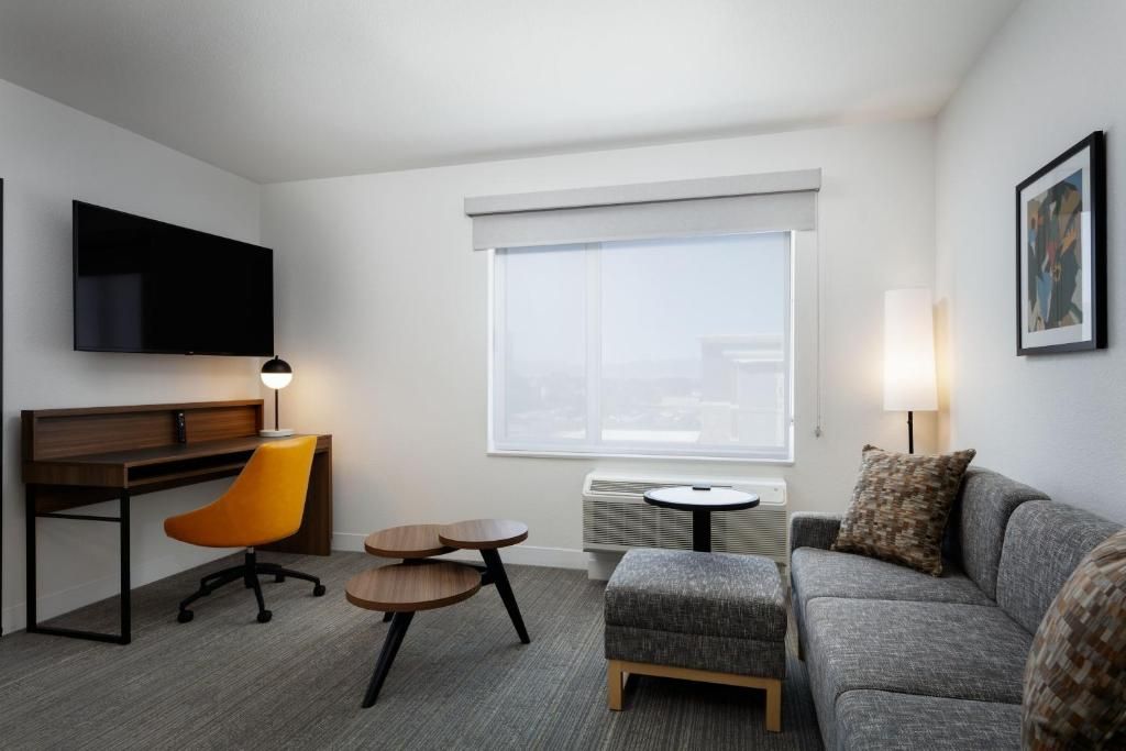 A hotel room with a flat-screen TV, a desk with a chair, a grey sofa, a small round table, and a large window with blinds.