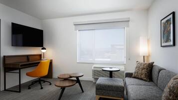 A hotel room with a flat-screen TV, a desk with a chair, a grey sofa, a small round table, and a large window with blinds.