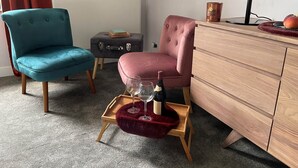 Una habitació amb una butaca color aguamarina, una butaca rosa, un armari de fusta i una taula petita amb un coixí vermell que sostenen una ampolla de vi i gots.