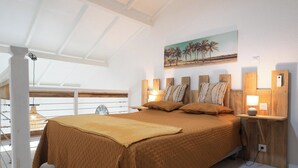 Une chambre avec un grand lit, une tête de lit en bois, deux tables de chevet avec des lampes, un tableau au mur et un balcon avec une balustrade.