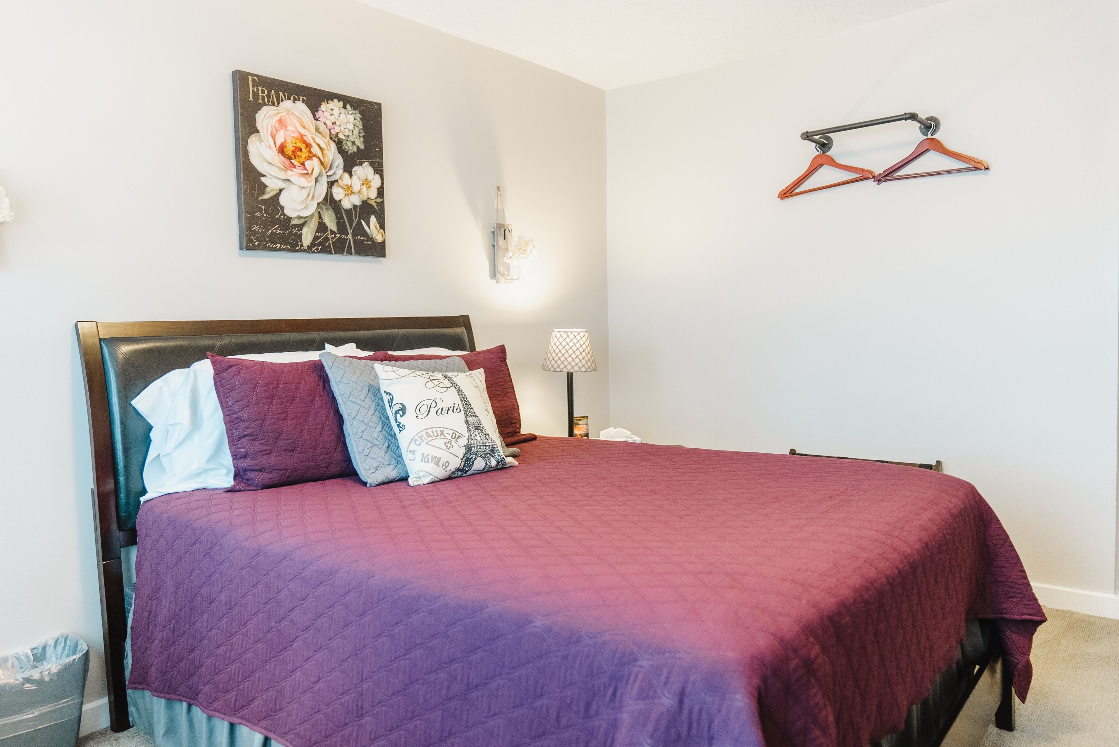 Una cama bien tendida con un edredón morado, una lámpara de pared y un cuadro decorativo de flores.
