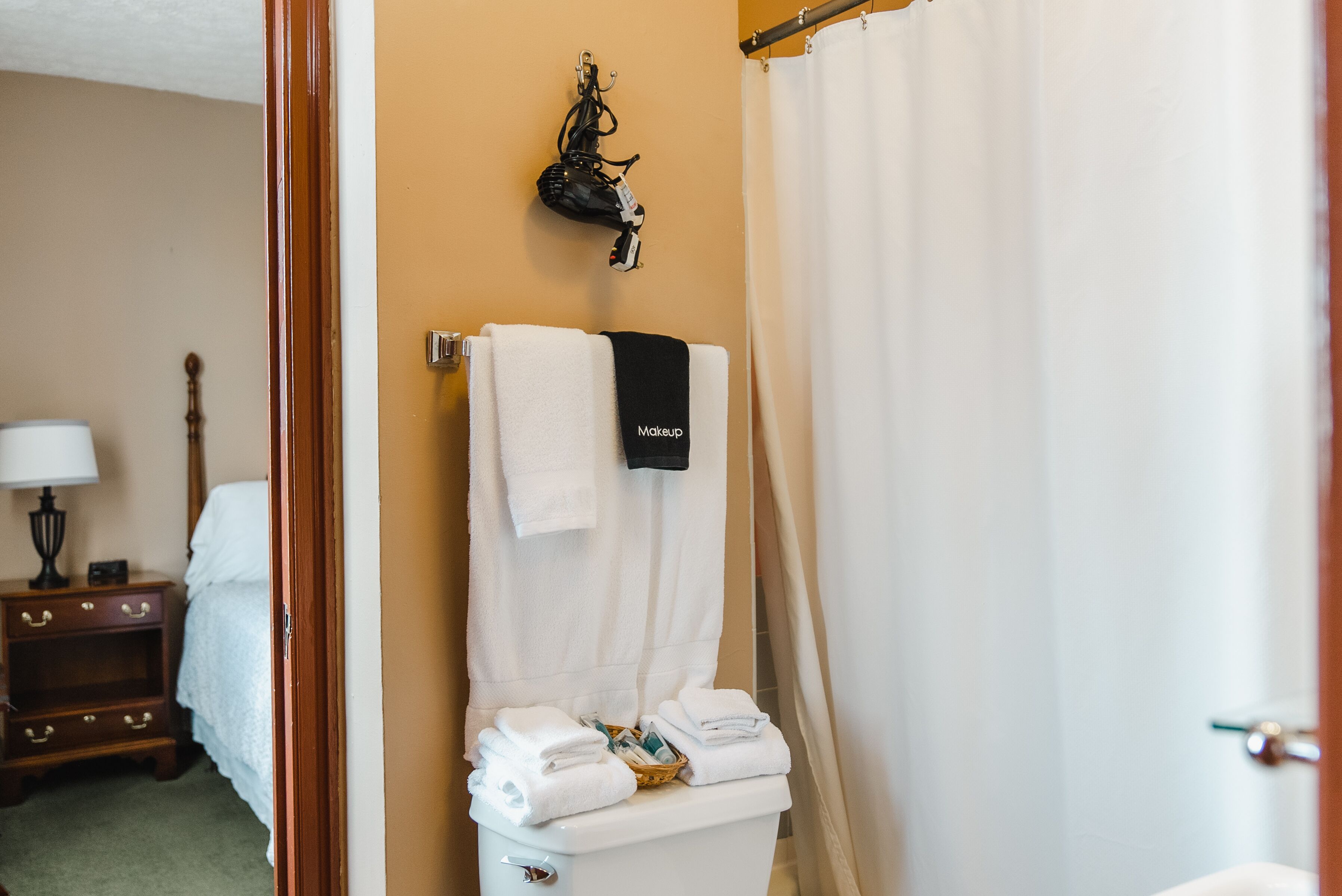 A bathroom with a toilet, a towel rack with a black towel, a basket of folded towels, and a wooden nightstand with a lamp.