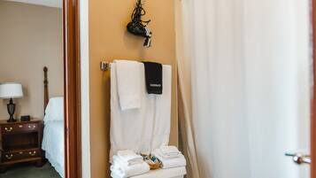 A bathroom with a toilet, a towel rack with a black towel, a basket of folded towels, and a wooden nightstand with a lamp.