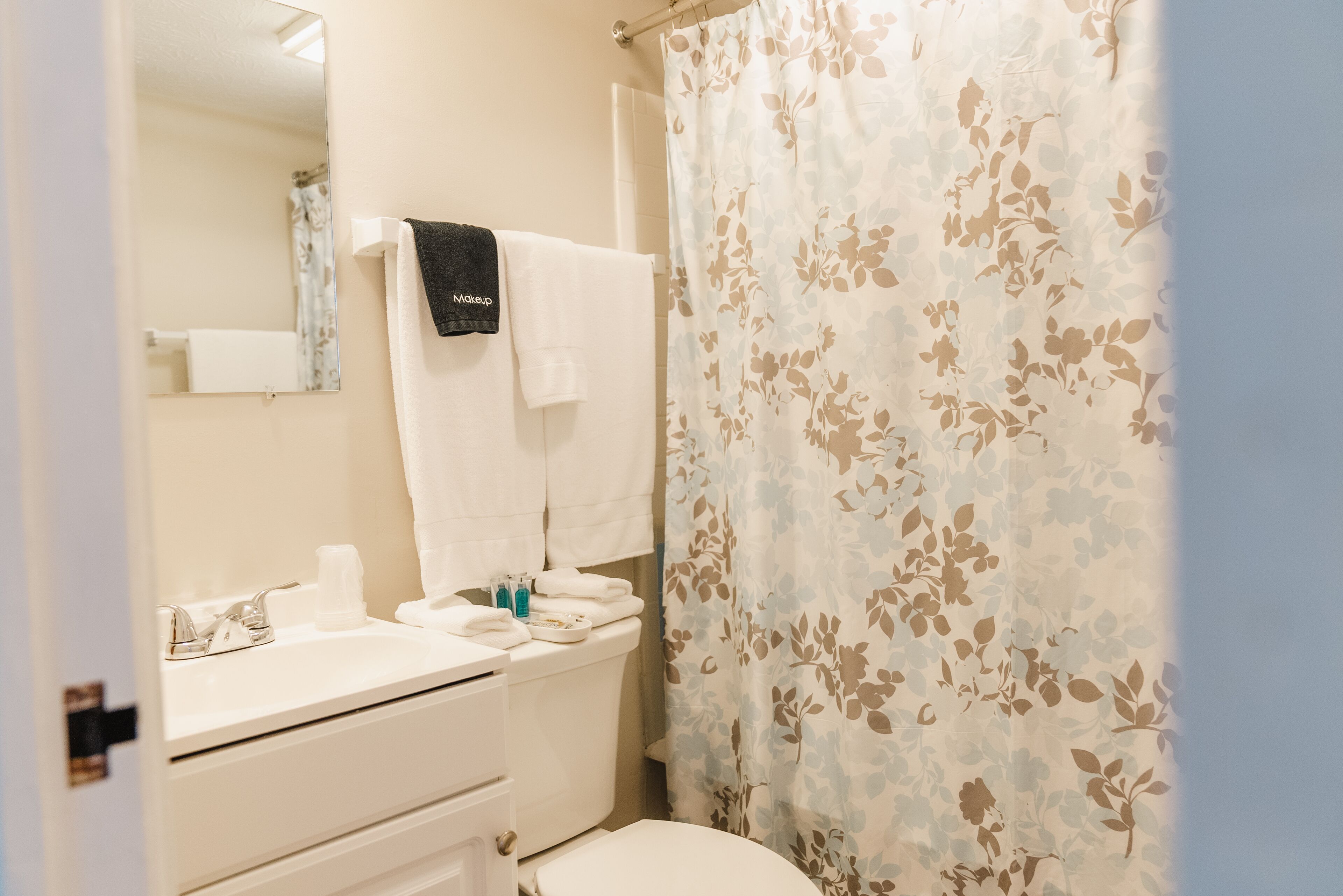 Een badkamer met toilet, wastafel en spiegel. Een handdoek hangt aan de muur en er is een douchegordijn met bloemenprint.