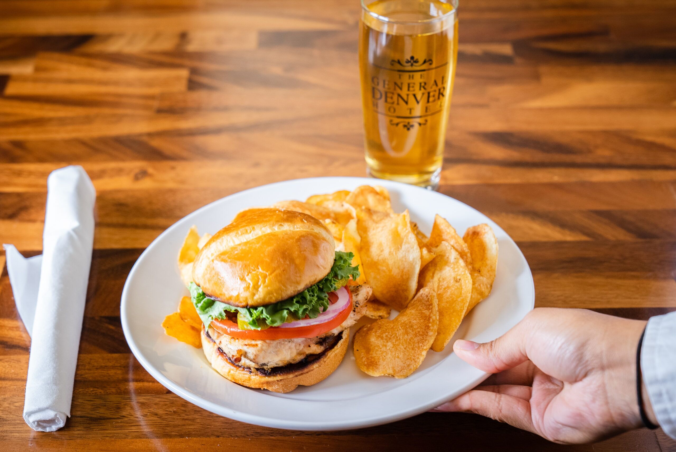 A plate with a burger and chips, a glass of beer, and a napkin on a wooden table.
