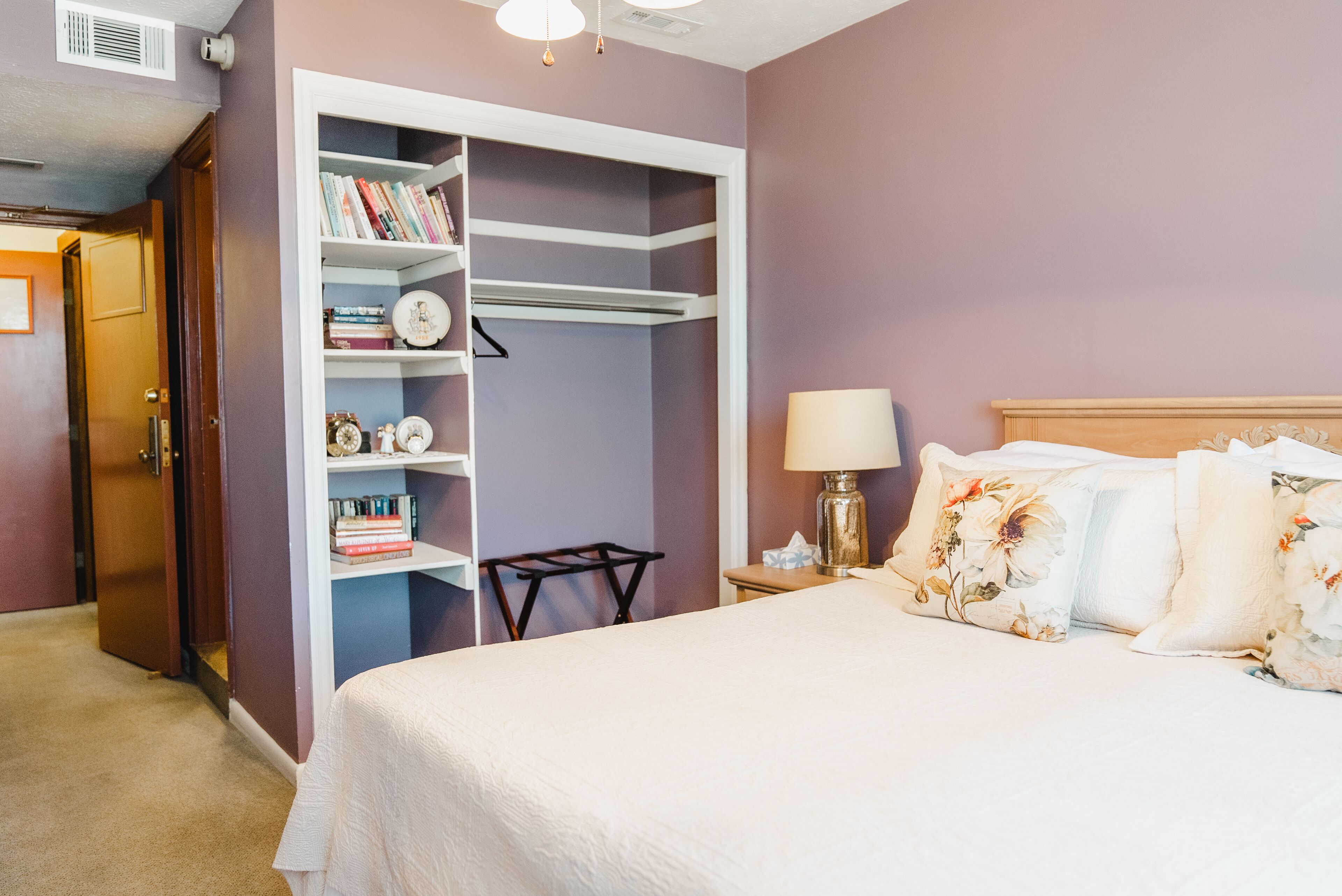A bedroom with a bed, a nightstand, a lamp, a bookshelf, and a door to a closet.