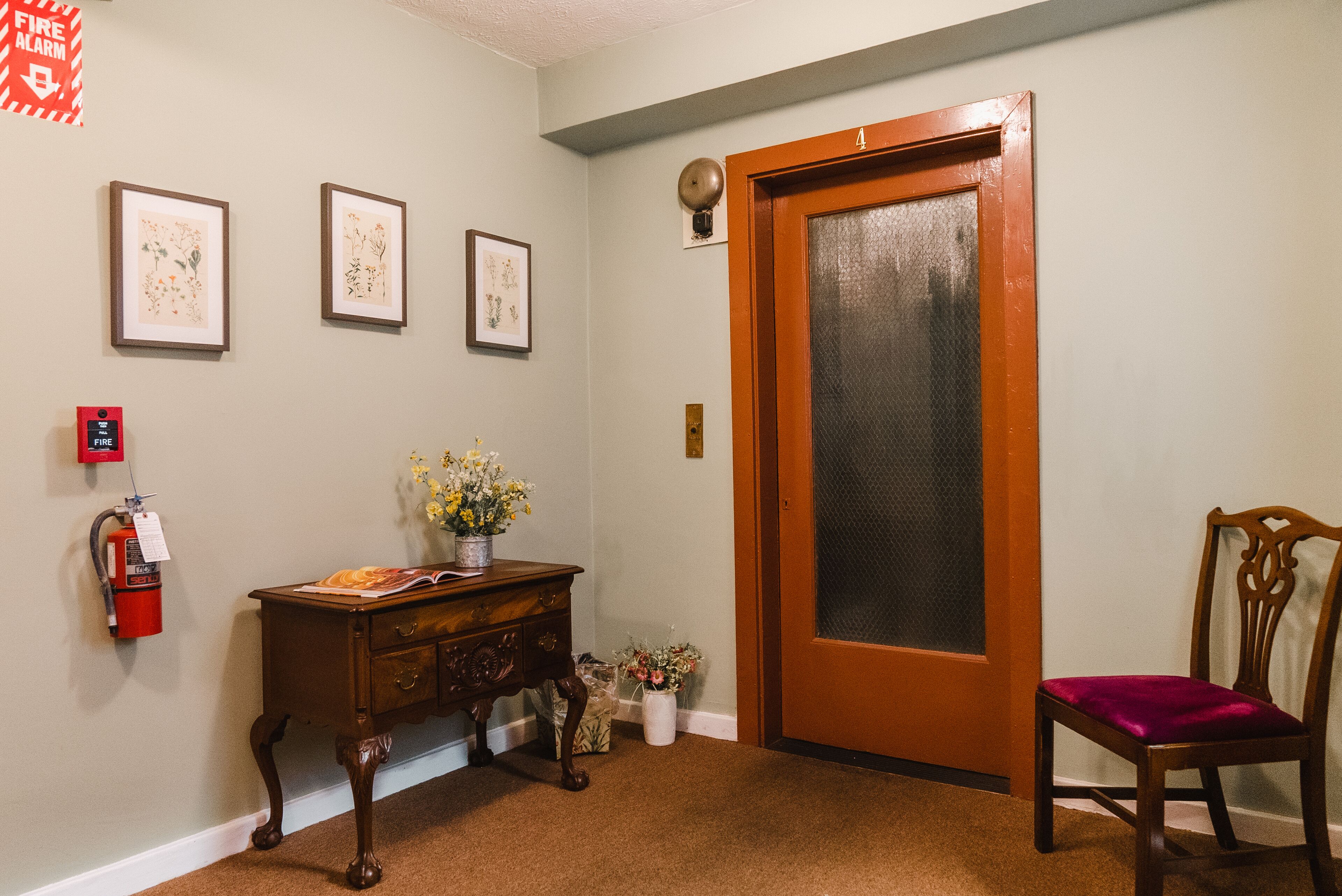 A hallway with a wooden door, a fire alarm, a desk with a vase of flowers, and a chair.