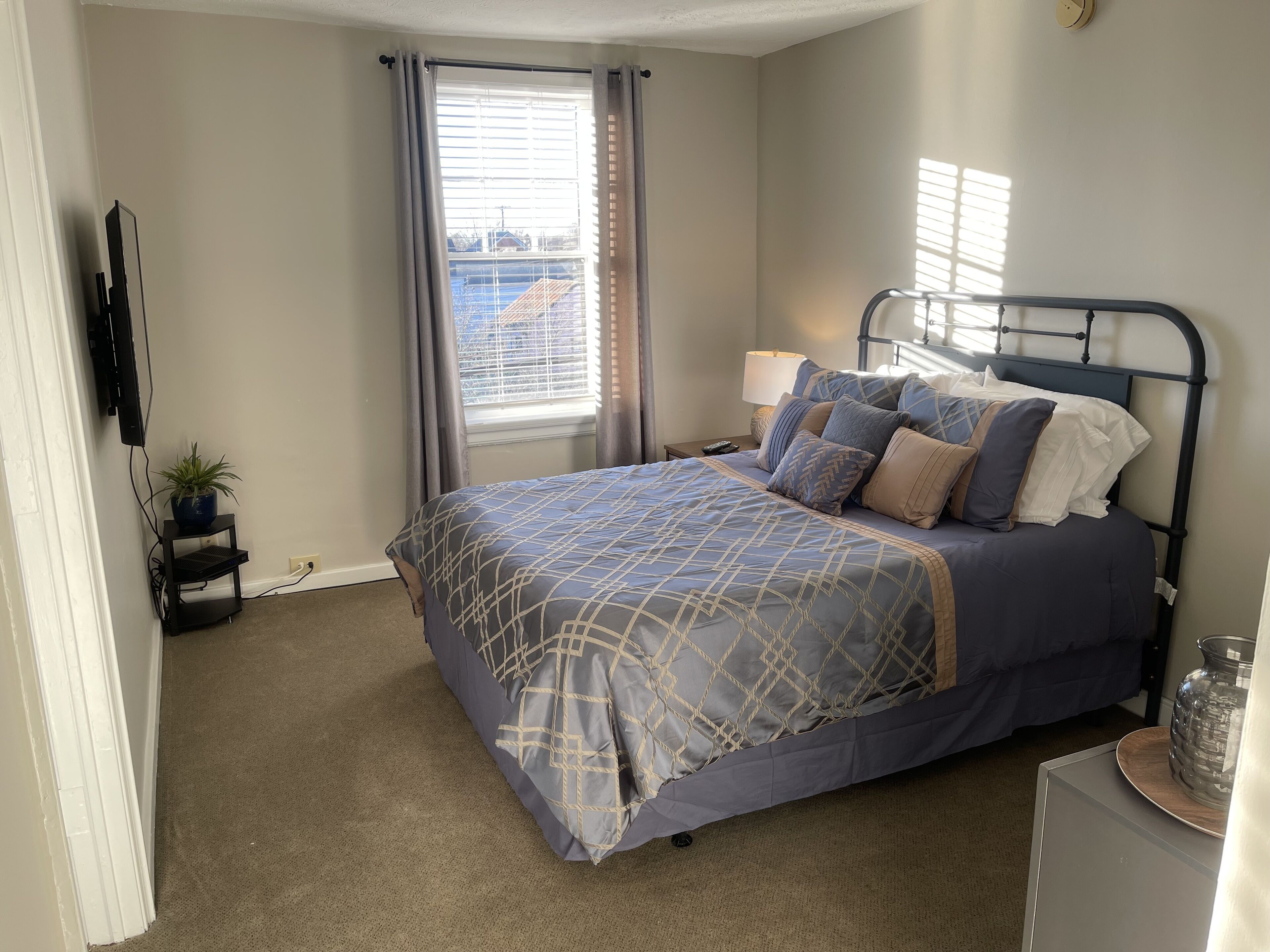 A bedroom with a bed, a bedside table, a lamp, a potted plant, and a window with curtains.
