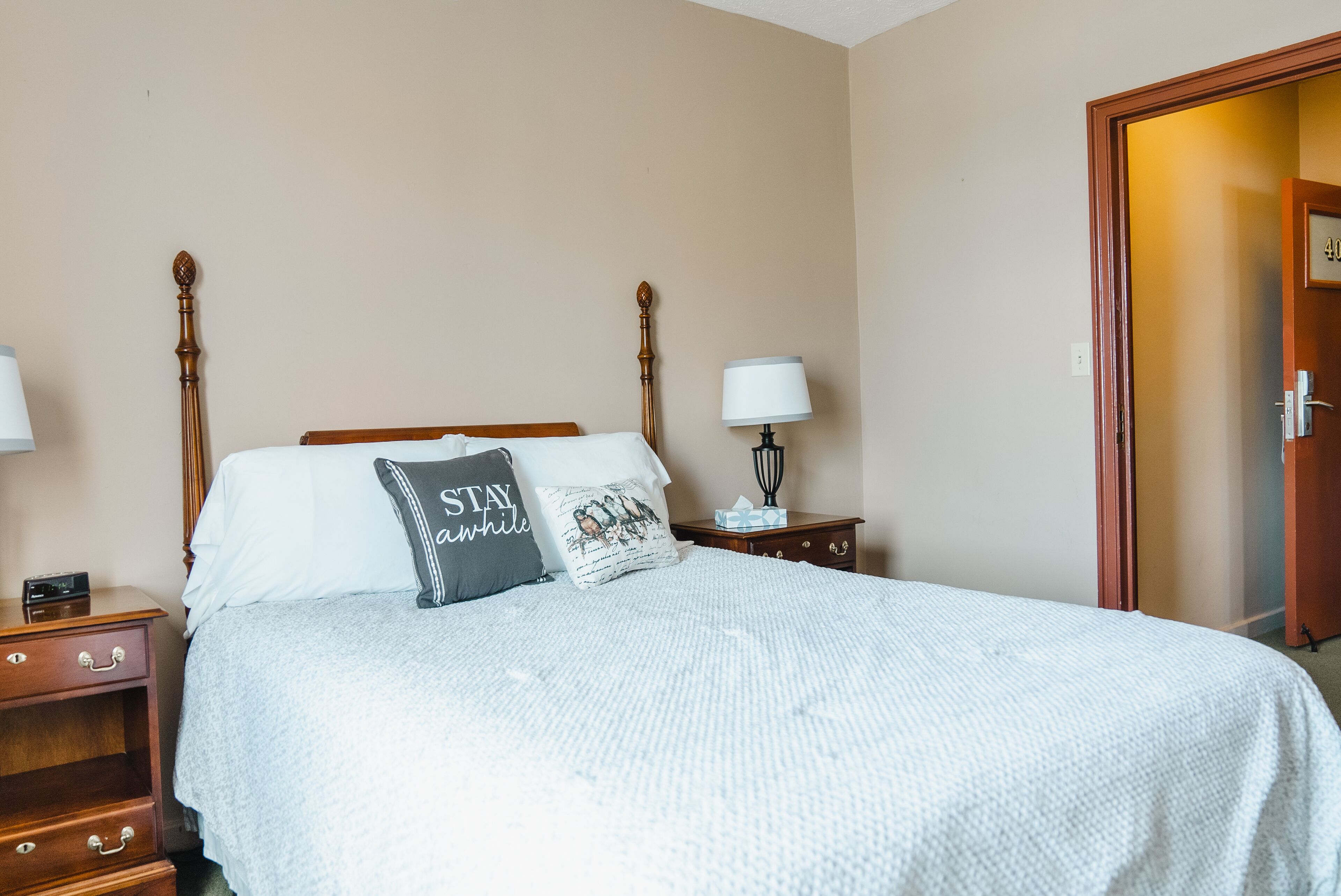 A neatly made bed with a white comforter and two pillows, one with a 'Stay Awake' design, in a room with beige walls and a wooden headboard.
