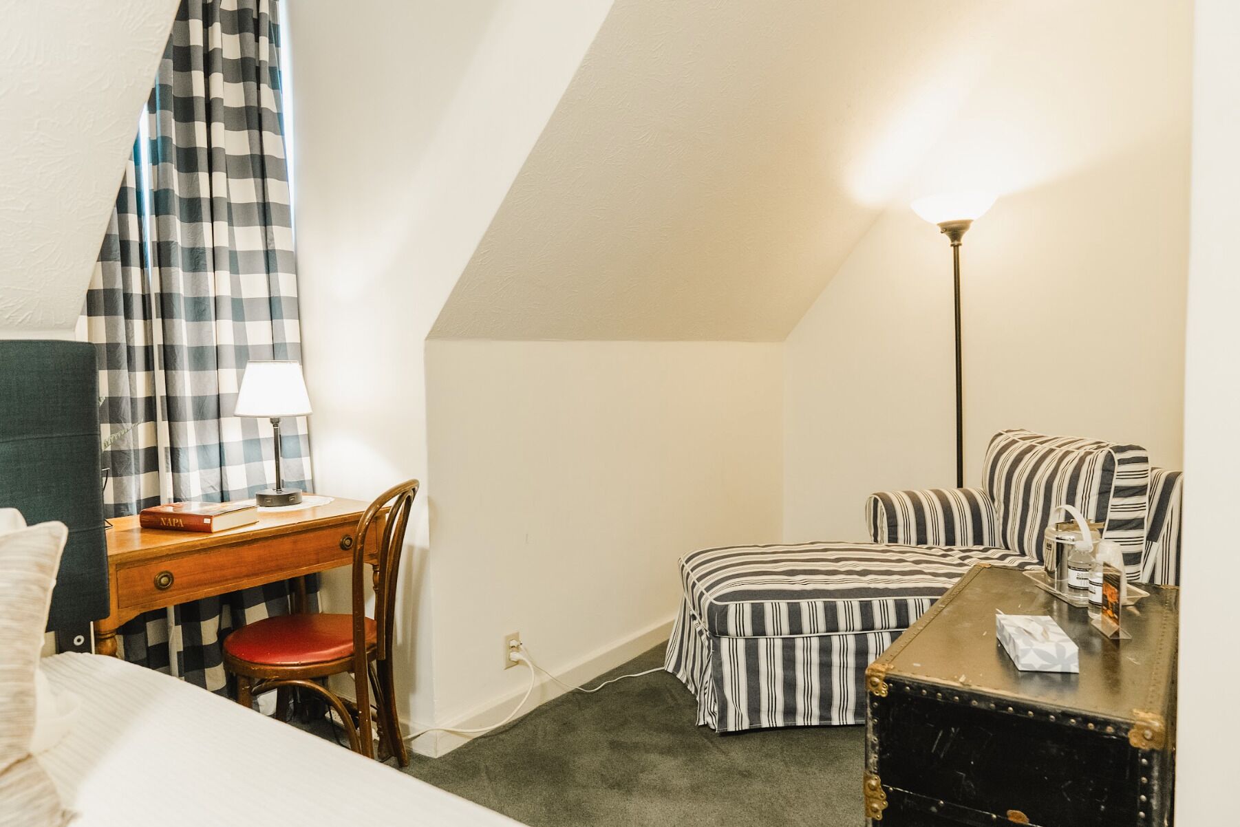 Ein kleiner, gemütlicher Raum mit einem Bett, einem Schreibtisch mit Stuhl, einem gestreiften Sessel, einem kleinen Tisch und einer Lampe.