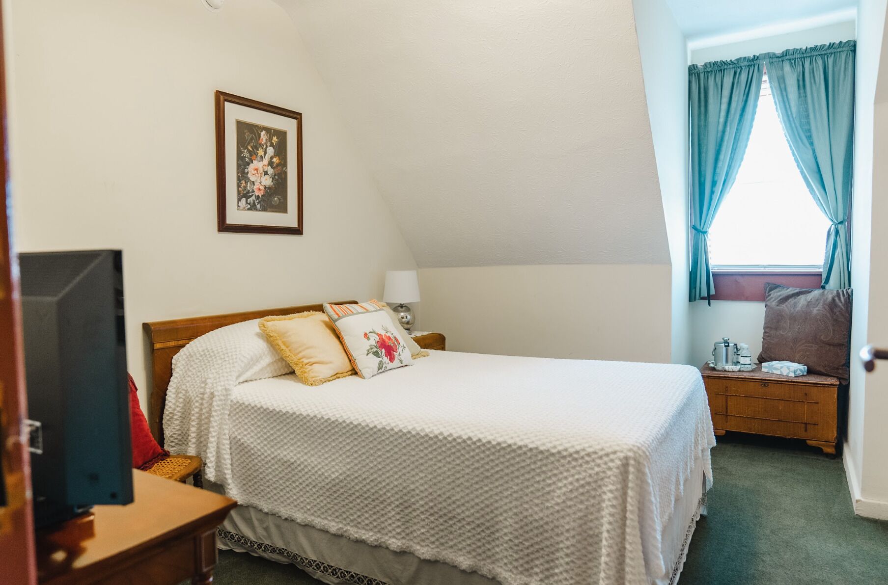 Une chambre bien rangée, avec un lit, une table de chevet sur laquelle se trouve une lampe, un tableau encadré au mur et une fenêtre avec des rideaux.