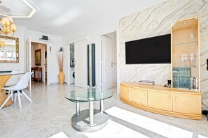 Un salon moderne avec une table en verre, un téléviseur fixé sur un mur en marbre et un meuble en bois doté d’étagères en verre.