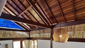 Un salón moderno con techo de madera, sofá gris con cojines amarillos, lámpara colgante de fibras tejidas y vista a una terraza exterior a través de un ventanal.
