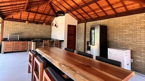 Un comedor amplio con una larga mesa de madera y bancos, una pared de ladrillo y una zona de cocina al fondo.