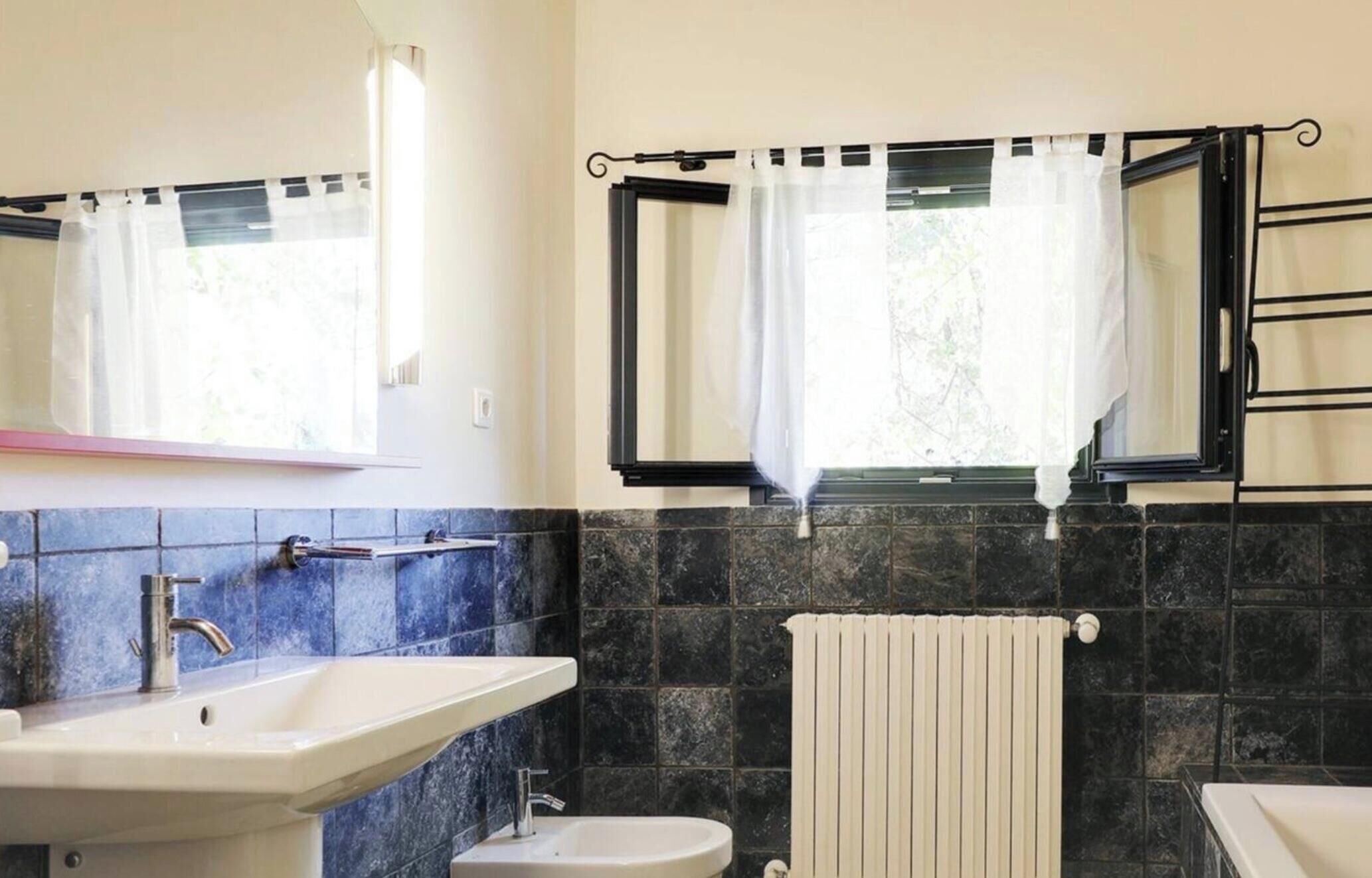 Un baño con lavamanos, espejo, ventana con cortinas y un perchero para toallas.