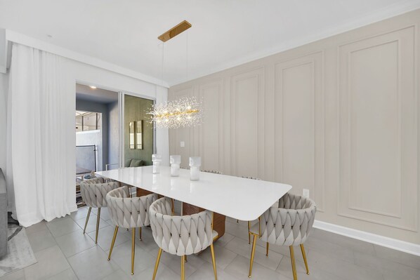 A modern dining room with a white table, six tufted chairs, and a unique chandelier.