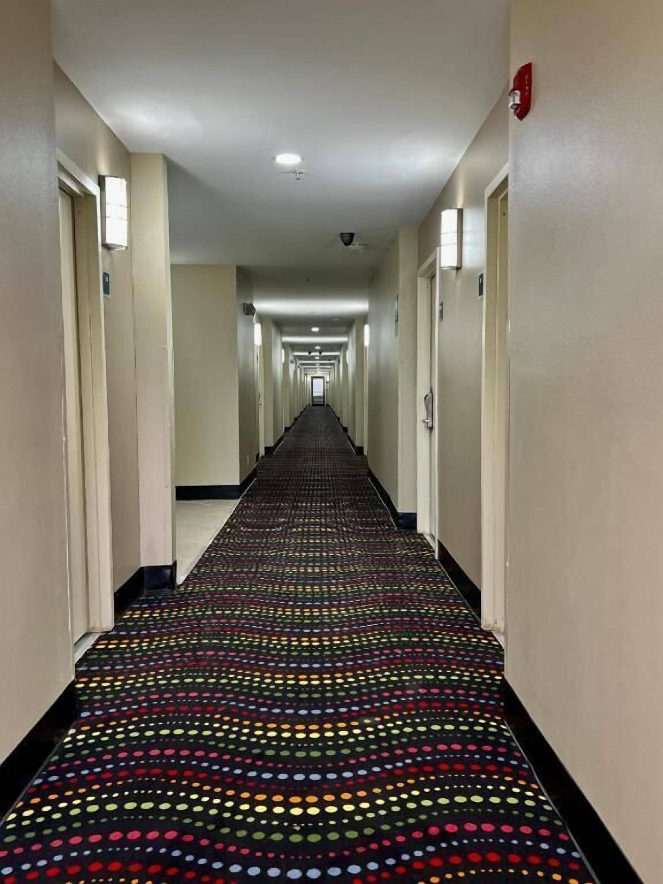 Un pasillo de hotel con alfombra estampada, puertas a ambos lados y un detector de incendios en la pared derecha.