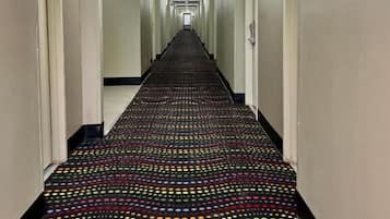 Un pasillo de hotel con alfombra estampada, puertas a ambos lados y un detector de incendios en la pared derecha.