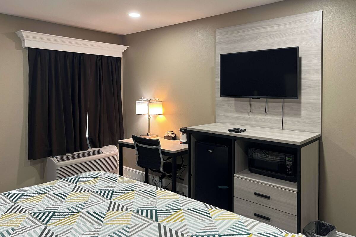 A hotel room with a bed, a desk with a chair, a television mounted on the wall, and a heater.