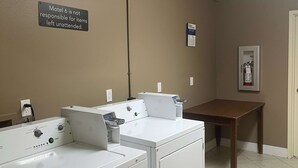 Una sala de bugaderia amb dues rentadores i assecadores blanques, un cartell que diu 'Motel 6 no es fa responsable d'articles deixats sense supervisió', i un extintor a la paret.
