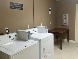 A laundry room with two white washers and dryers, a sign stating 'Motel 6 is not responsible for items left unattended', and a fire extinguisher on the wall.