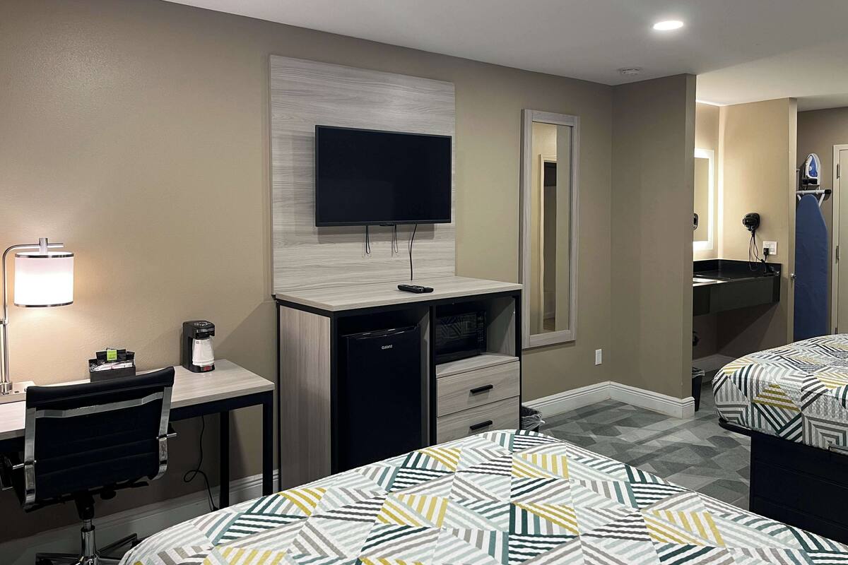 A hotel room with a desk, a flat-screen TV, a mirror, a bed with patterned bedding, and a small kitchenette.
