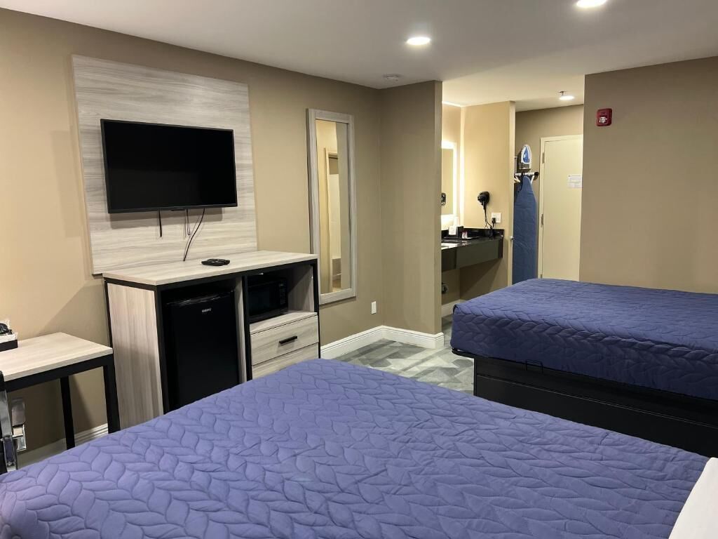 A hotel room with a flat-screen TV mounted on the wall, a desk with a computer, a small table, two beds with blue bedding, and a bathroom visible through an open door.