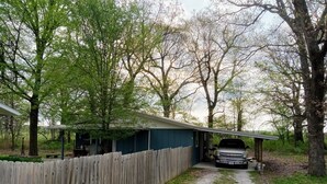 A covered garage with a vehicle parked inside, surrounded by trees and a wooden fence.