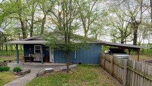 A small blue shed with a covered porch, a barbecue grill, and a vehicle parked outside.