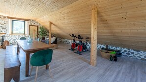 Un plafond en bois, des murs en pierre, une table à manger, des chaises, un fauteuil vert, deux vélos d’appartement et un panier rempli de poids.