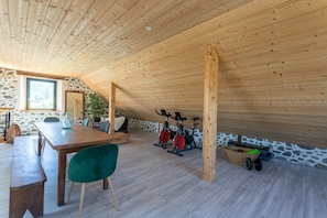 A wooden ceiling, stone walls, a dining table, chairs, a green chair, two exercise bikes, and a basket with weights.