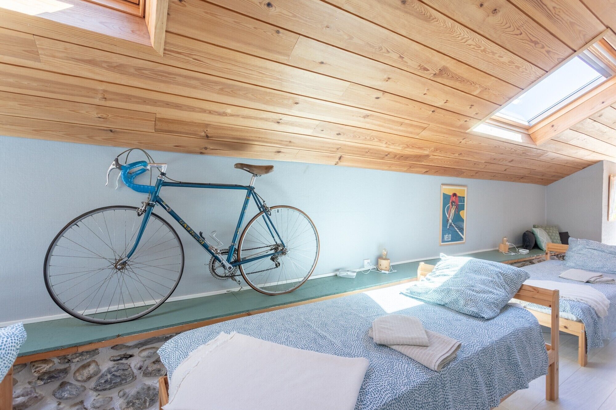 A room with a wooden ceiling, a bicycle, two beds with patterned bedding, a stone floor, and a poster on the wall.