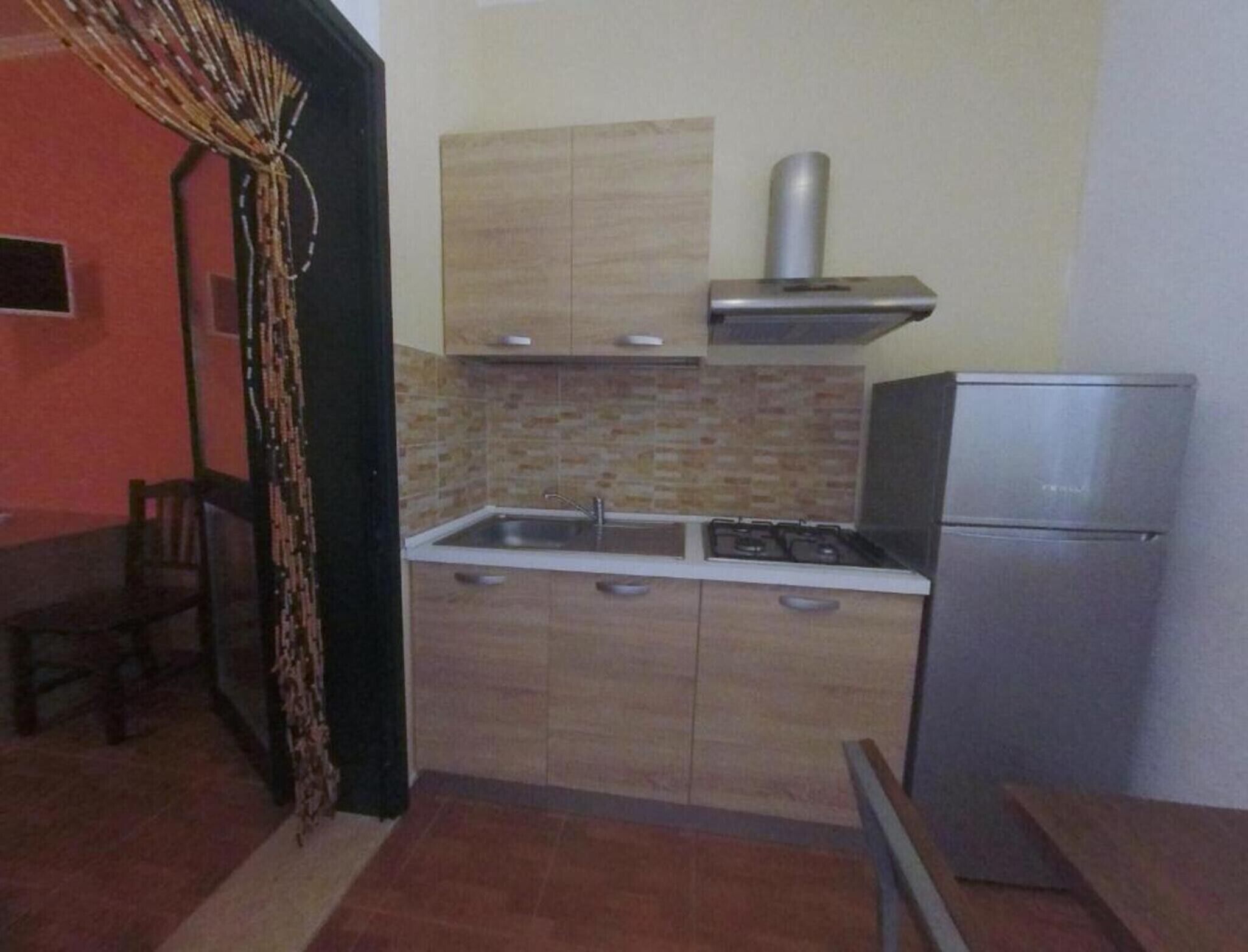 Een kleine keuken met een fornuis, spoelbak en koelkast, houten kastjes en een betegelde spatwand.