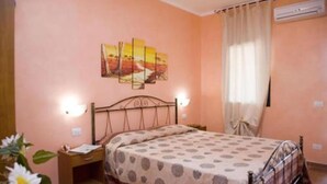 Un dormitorio con cama, mesita de noche, lámpara de pared, ventana con cortinas y un cuadro en la pared.