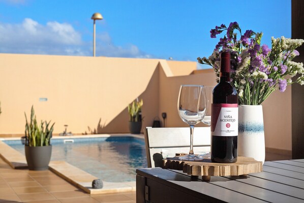 A bottle of wine, a glass, and a vase of flowers on a wooden table by a poolside.