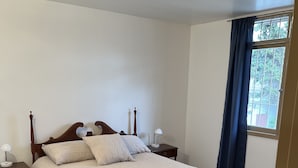 Ein Schlafzimmer mit einem hölzernen Bett, einem Nachttisch, einer Lampe und Blick ins Freie durch ein Fenster mit Vorhängen.